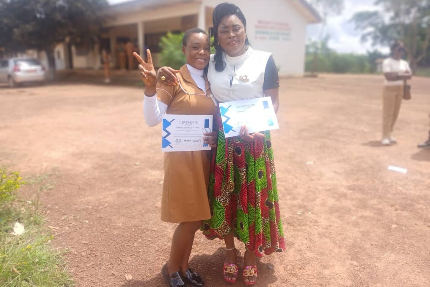 Two women holding certificates and posing outdoors in front of a building, one making a peace sign.