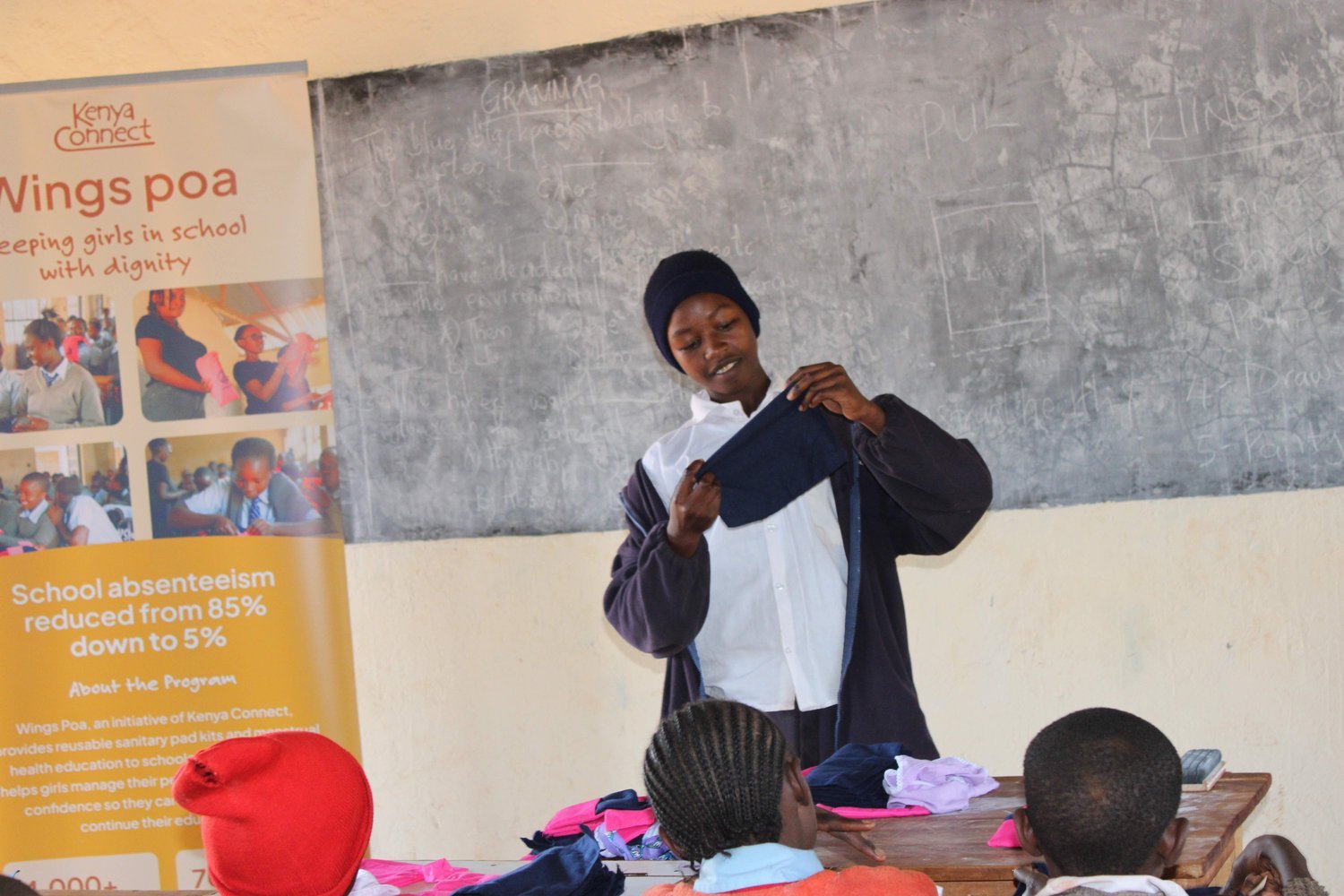 A woman holds up a dark blue garment in a classroom beside a Kenya Connect "Wings Poa" programme banner.