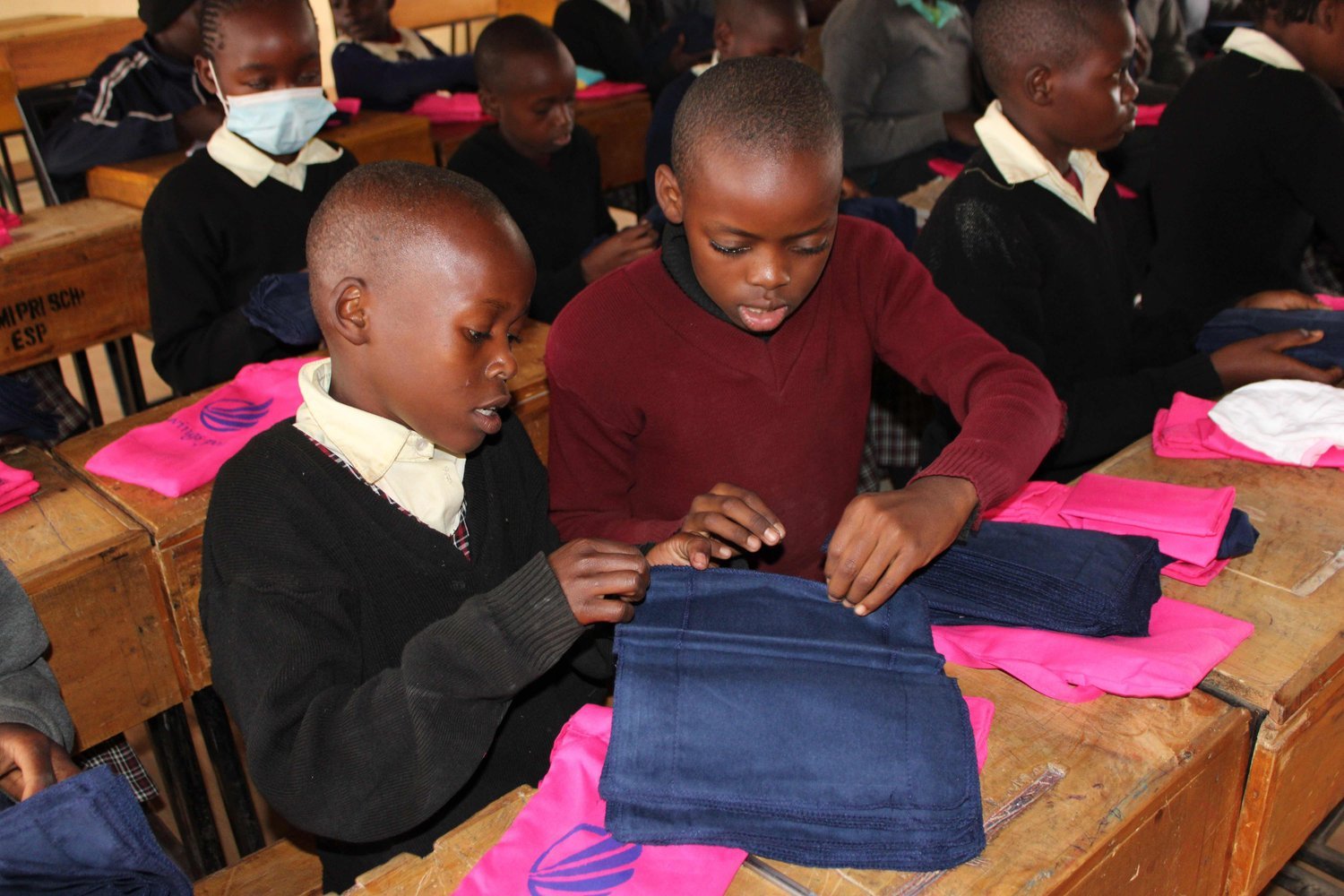 Children in school uniforms examining folded blue and pink fabric items at wooden desks in a classroom.
