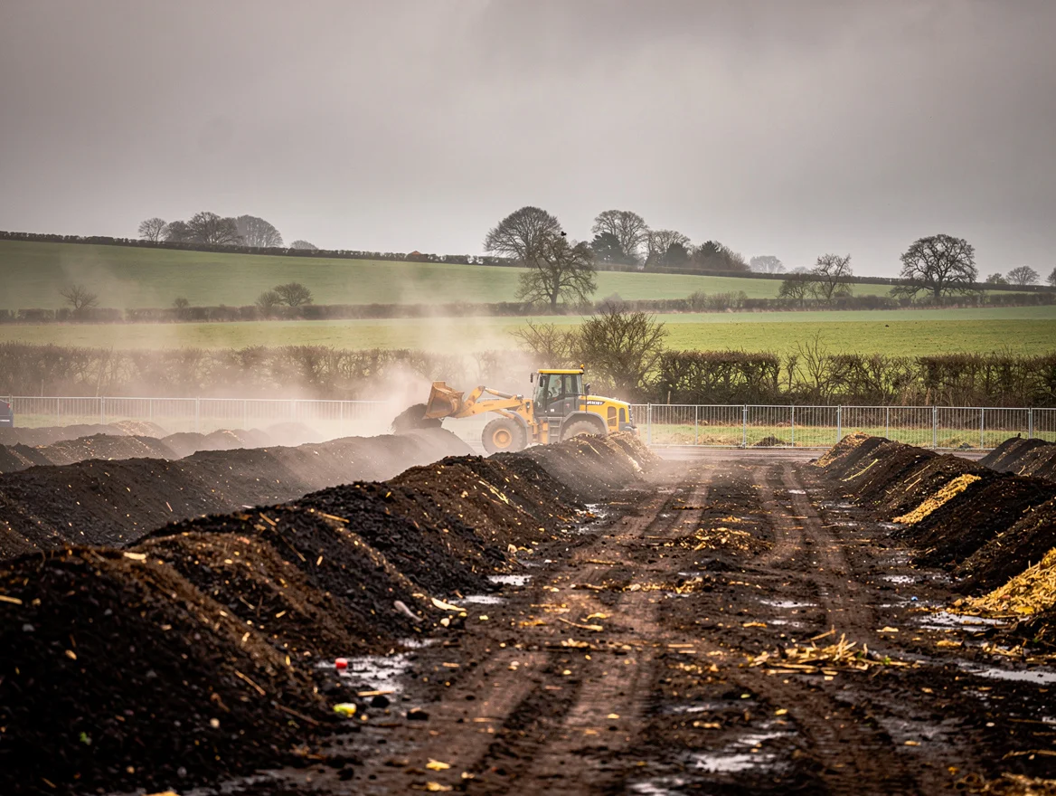 Industrial composting facility in the UK with steaming windrows of organic material being turned by a front loader under overcast skies