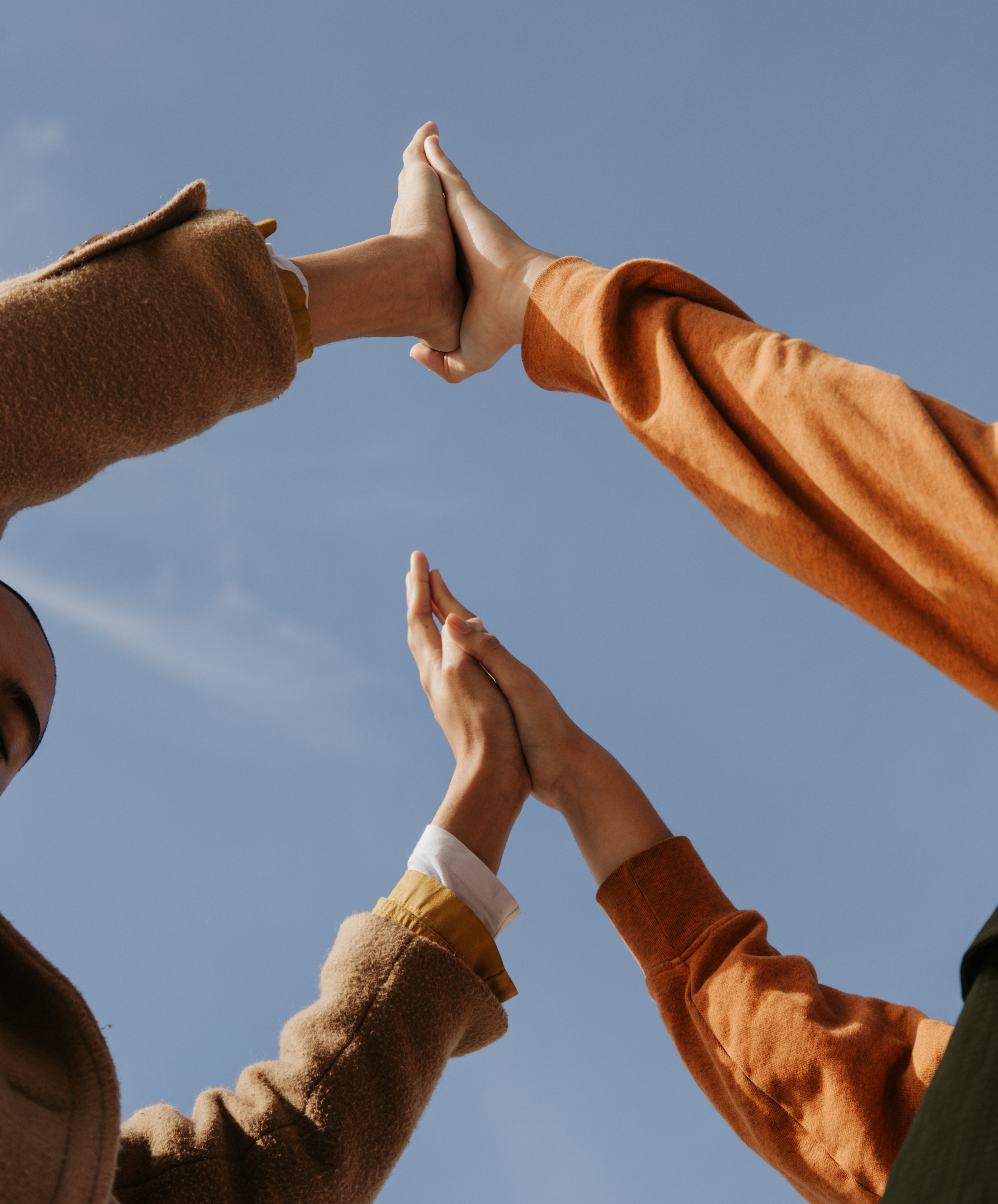 Four hands wearing autumn-colored clothing meet in two high-five gestures against a clear blue sky, symbolizing teamwork, unity, or connection.