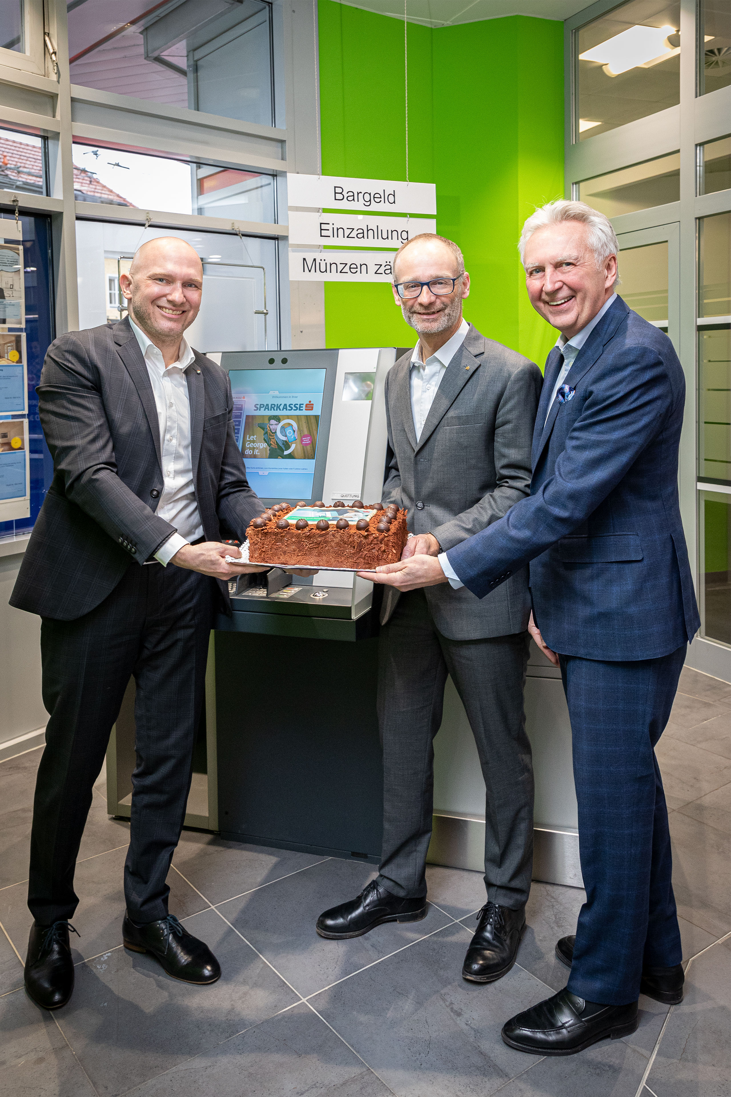 Three men presenting the festive cake for the party in front of the jubilee ATM of the evo series.