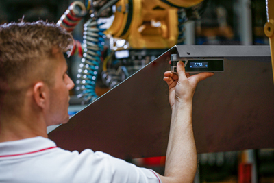 Employee of a production company measures the angle of a large sheet metal part. The angle measuring device is held upwards and shows the measured angle on the display.