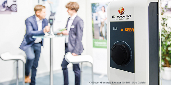 Eine weiße KEBA Wallbox mit einem aufgedruckten Logo der Messe „E-world energy & water“ am KEBA Messestand - im Hintergrund unterhalten sich zwei Männer an einem Stehtisch.