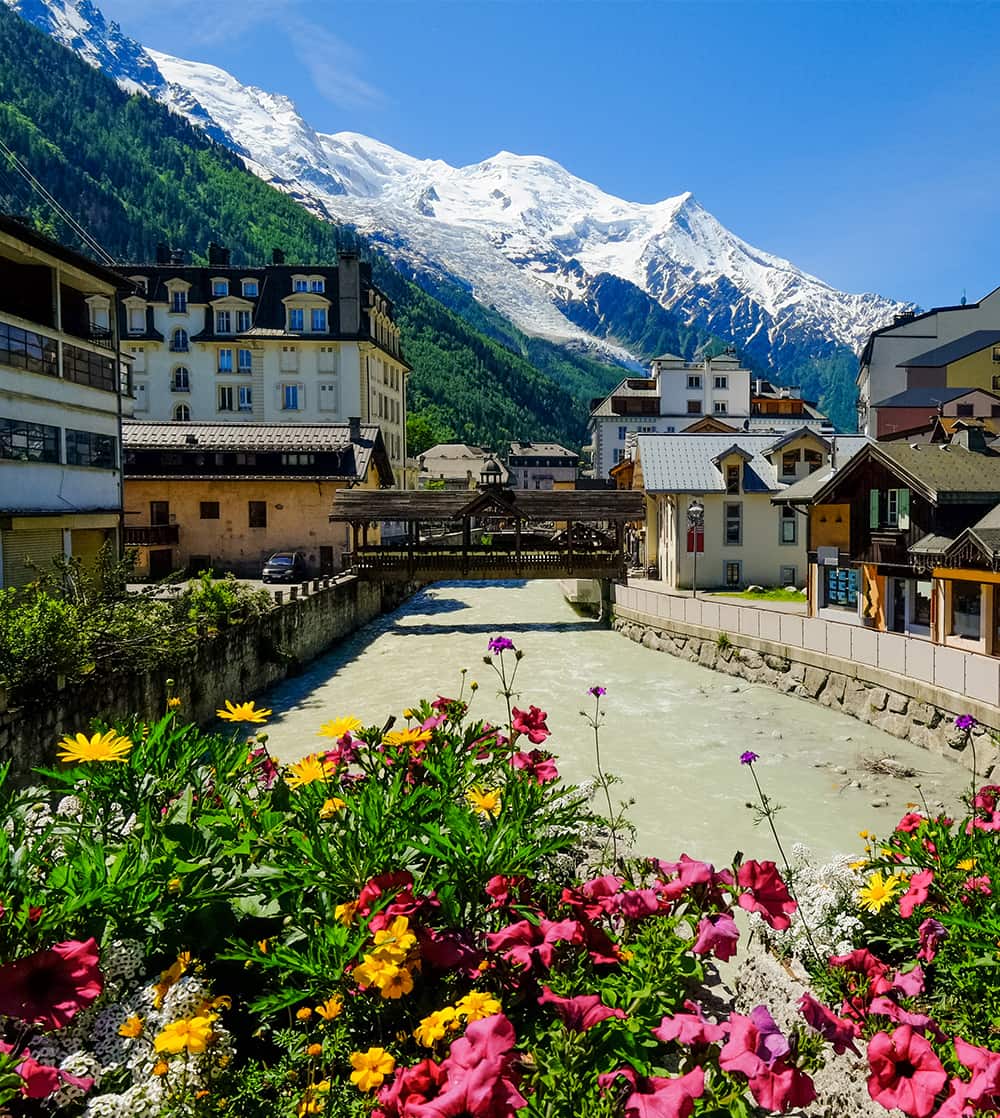 A river running through a medieval town with snow-capped mountains in the background
