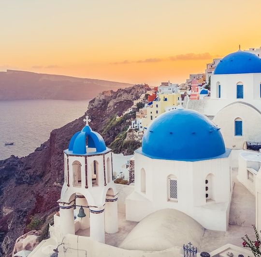 White buildings with blue domes in Santorini next to colorful homes overlooking a cliff during a sunset