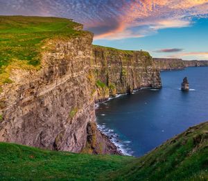 A large rocky cliff overlooking the ocean with grass at the top of the cliff during a cloudy sunset