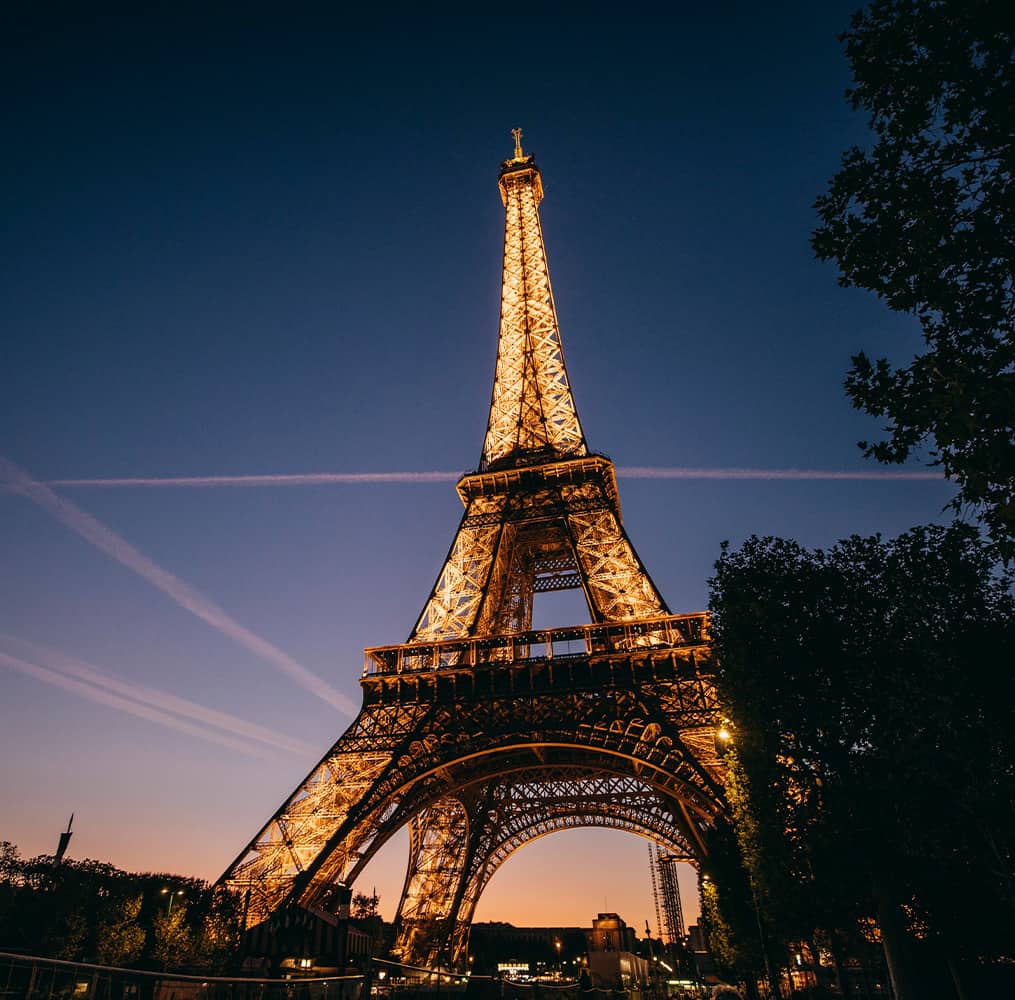 The Eiffel Tower lit up at night with several pink airplane contrails crisscrossing in the sky