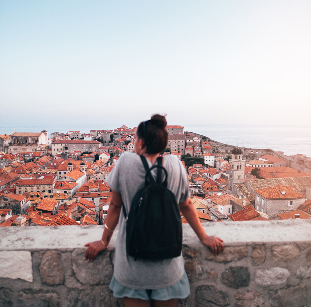 A person wearing a backpack facing away from the camera and looking over an aerial view of a city with many red roofs