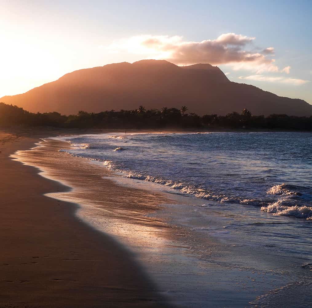 A beach with waves and a mountain in the background at sunset