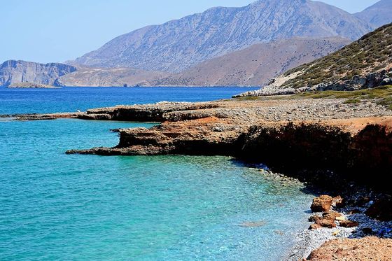 A rocky shore next to clear blue water with mountains in the background