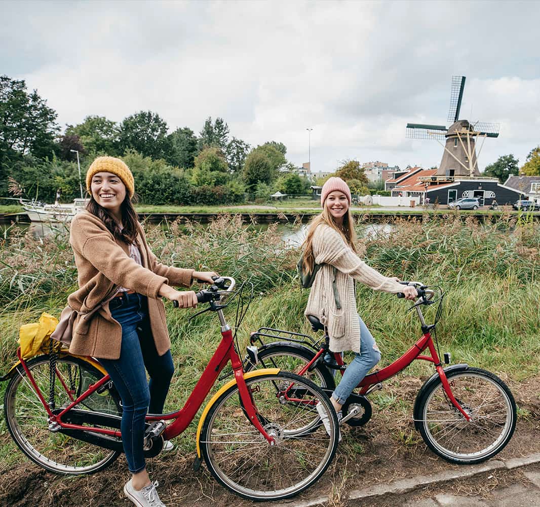 Two young women on bicycles on a grassy dirt road with a windmill in the background