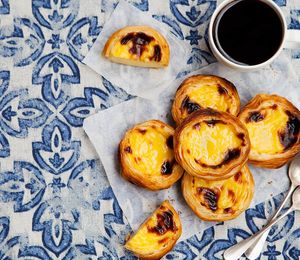 A tablecloth with a blue and white design covered with egg pastries and a mug of coffee