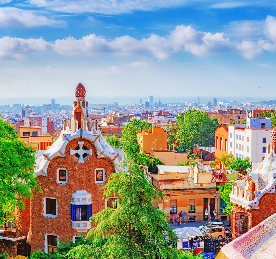 A view overlooking Park Guell which is a vibrant outdoor collection of gardens and architecture located in Barcelona. A red brick building with a white cross and white trim details is in the foreground with several trees. The city of Barcelona can be seen in the background with clouds scattering a blue sky.
