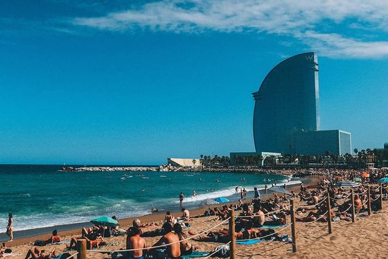 Many people lounging on towels on the sand next to the ocean with tall buildings in the background