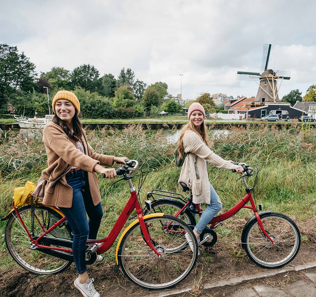Two young women on bicycles on a grassy dirt road with a windmill in the background