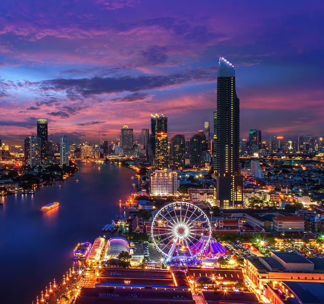 An aerial view of a city located next to a body of water during dusk with many buildings lit up with lights including a Ferris Wheel