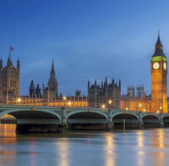 A bridge spanning a river and a large, ornate building with a clock tower behind it at dusk