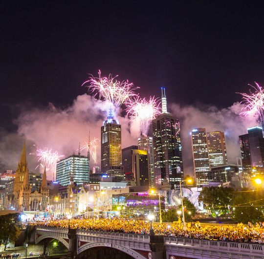 A city skyline at night with fireworks in the background and crowds of people in the streets in front