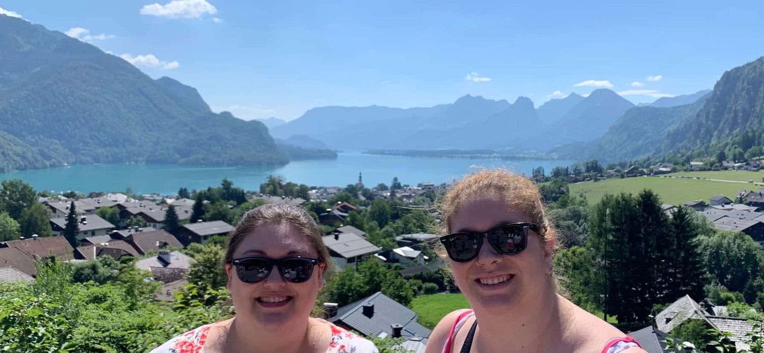 Two plus-size woman smiling with a scenic European mountain backdrop.