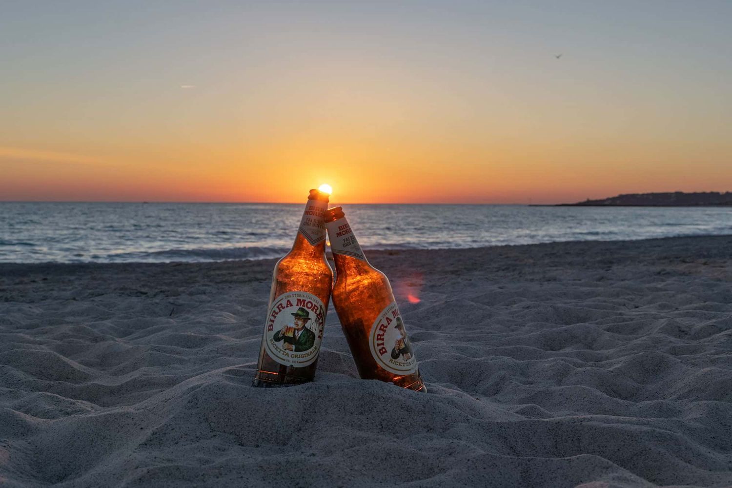 Two bottles of a local Italian beer, Birra Moretti, sitting in the sand on a beach.