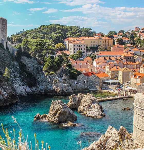 A cluster of homes with red roofs situated next to a forest and at the edge of a body of crystal clear blue water with some rocky formations scattered in the water