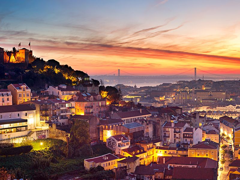 Yellow and orange sunset in Lisbon with bridge in background and lit-up buildings in foreground