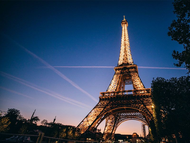 A view looking up at the Eiffel Tower lit up at night with contrails along the night sky in the background