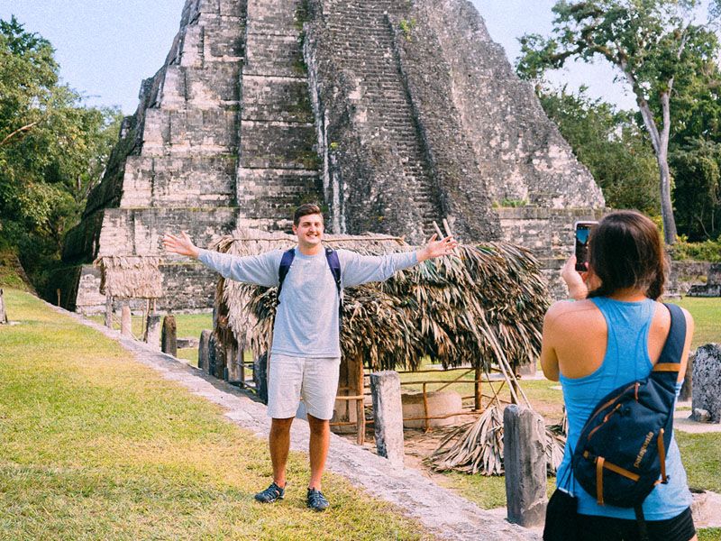 A woman taking a picture of a man with his arms spread wide in front of a stepped stone temple.