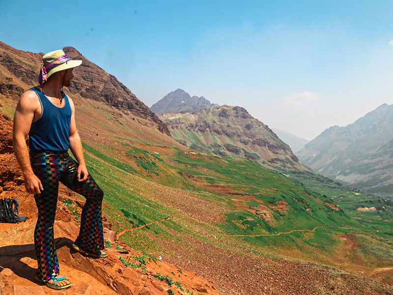 A man standing on a red dirt trail looking out at a view of mountains and valleys