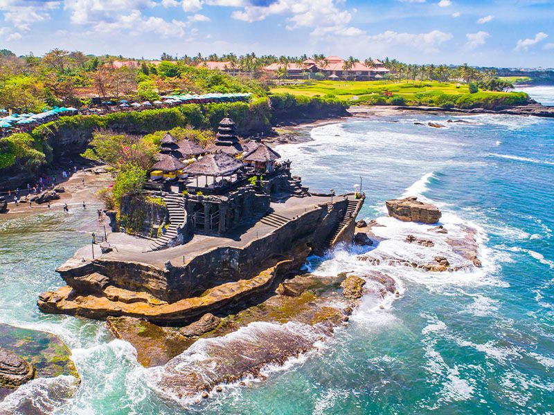 A temple on a rocky cliff in the ocean on a sunny day with umbrellas, buildings, and trees on the coast in the background.