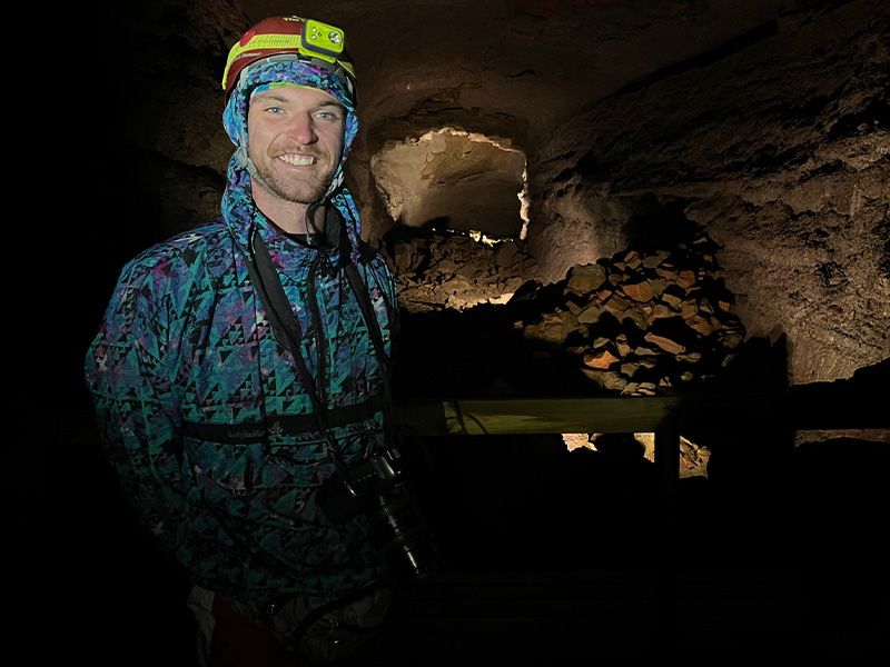 A man with a headlight and camera smiling for a photo in the inside of a rocky cave