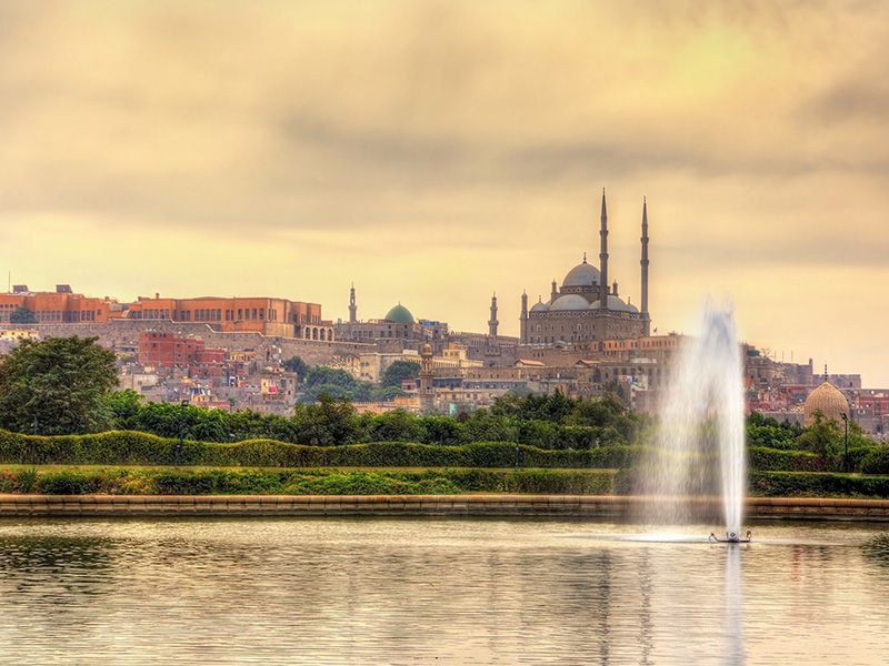 A lake and fountain with greenery, a city skyline, and mosque in the background.