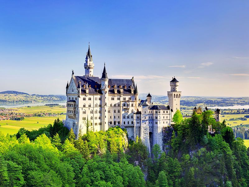 Large white castle perched on a tree-lined hill with green grass and blue sky in the background