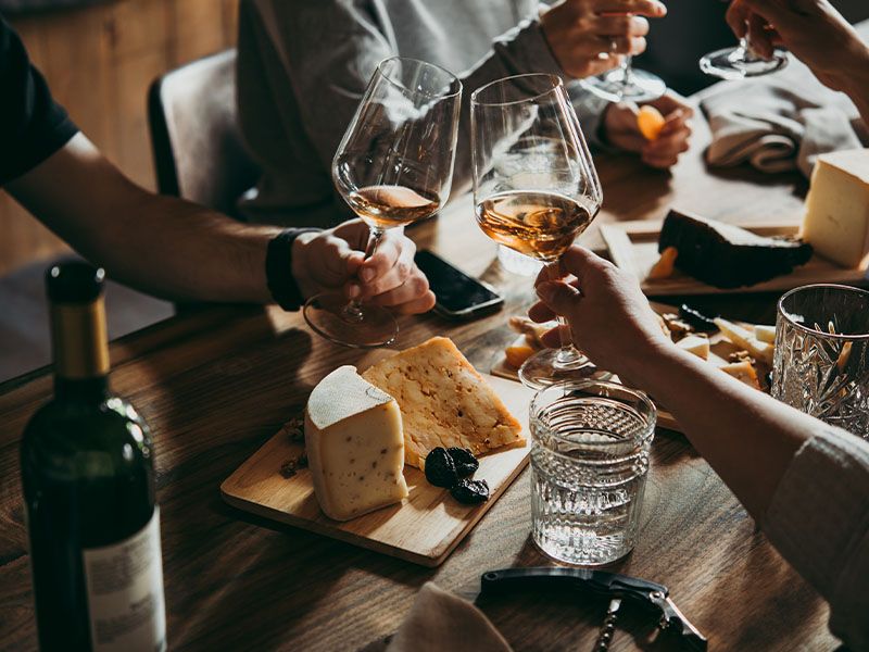 Two people's hands holding filled wine glasses about to clink their glasses together over a table covered with water glasses and a cheese plate