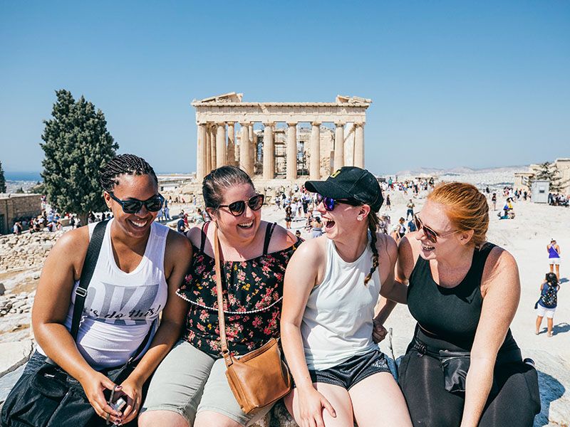 Four women smiling and sitting in front of Greek ruins with people milling in the background.