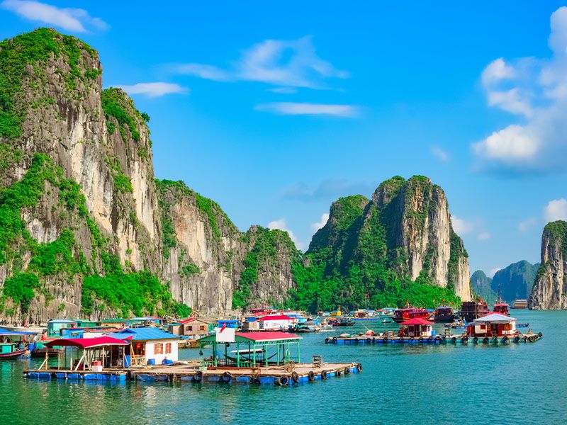 Colorful shacks floating on docks and boats in a bay with tall, greenery-covered rock formations in the background.