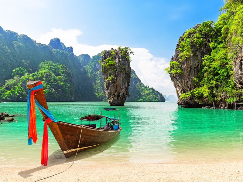 A traditional wooden Thai boat resting on the shore of a tropical bay with greenery-covered islands in the background.