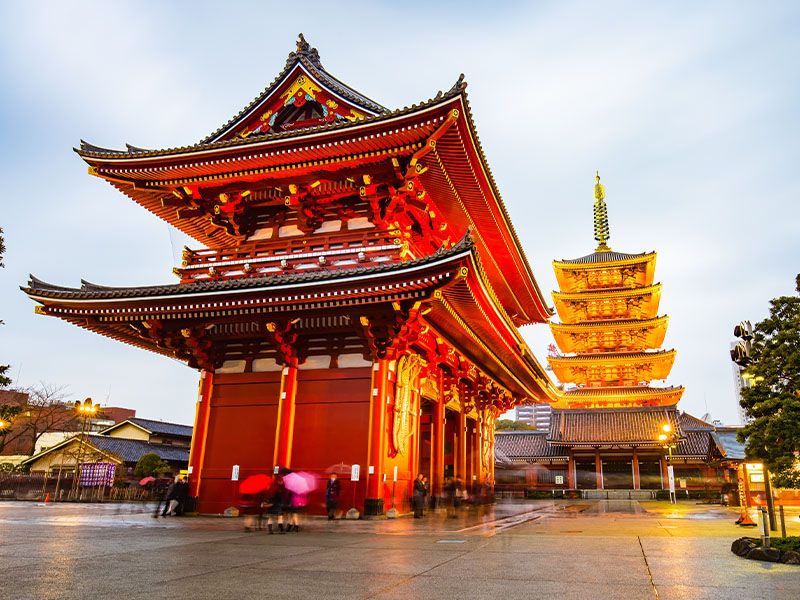 People with umbrellas standing at the base of a red Japanese pagoda with a yellow pagoda in the background.