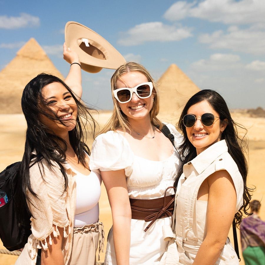 Three women smiling and standing in front of the Pyramids of Giza