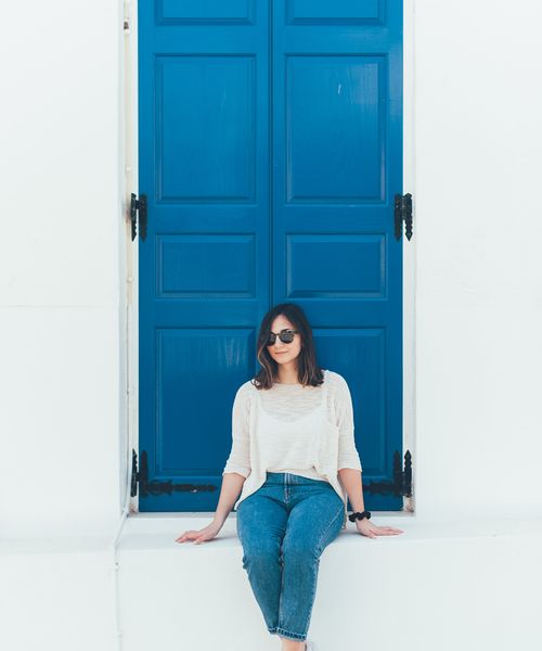 a woman sitting in front of a bright blue door on a traditional greek whitewashed building