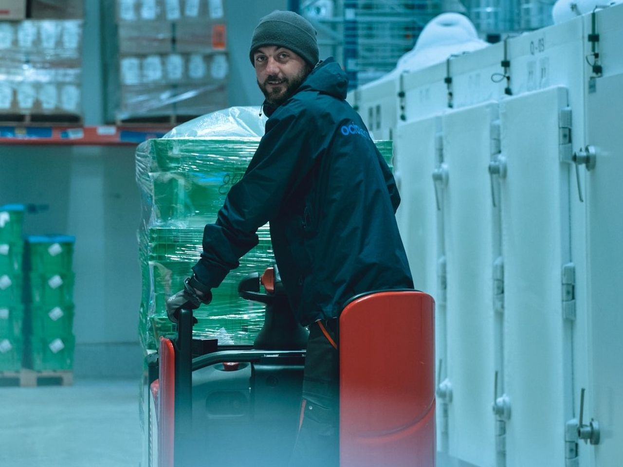 Un homme sur un transpalette regarde la caméra. Il porte un bonnet et une grosse veste.