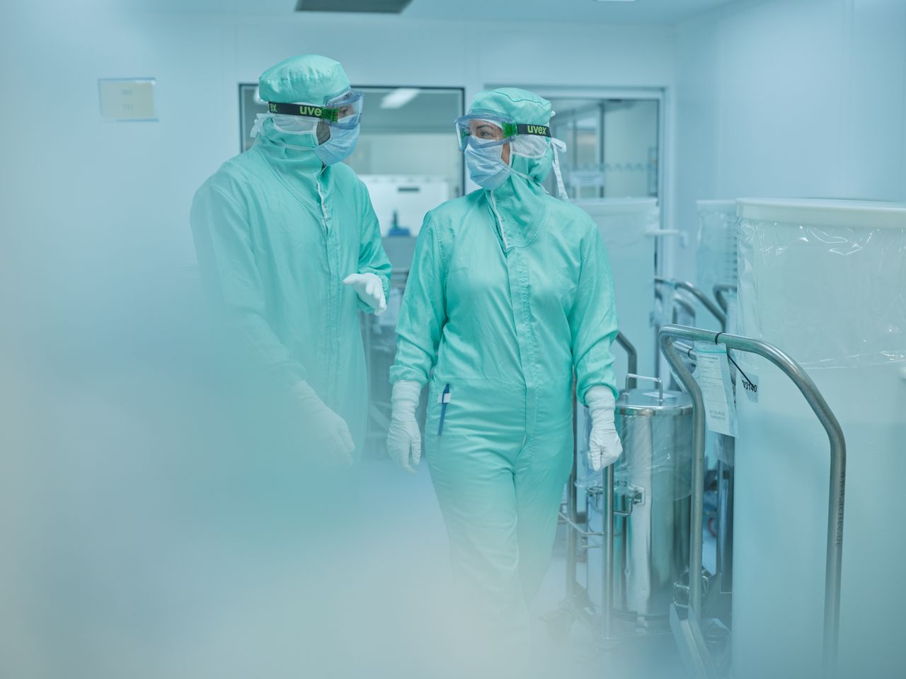 Une femme marche avec son collègue dans un couloir en zone de production. Ils sont couvert de la tête aux pieds avec leur tenue aseptique. Il discutent et on remarque leur sourire grâce à leurs yeux.