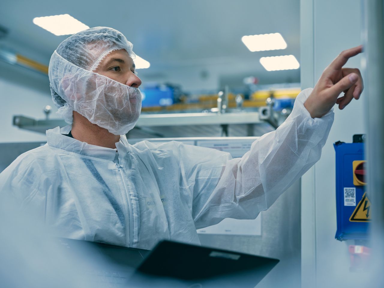 Un homme, de profil, portant une charlotte et un cache barbe. Il travaille sur un automate en zone de production.