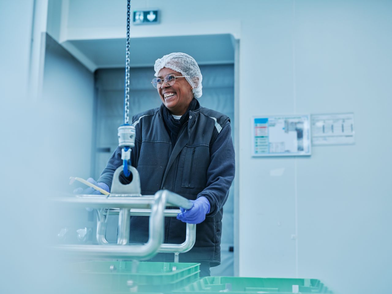 Une femme, très souriante, est en train porter une caisse de plasma avec un appareil d'aide au port de charge lourde. Elle porte une charlotte sur la tête et des gants violets.
