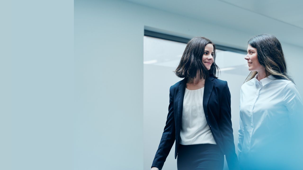 Zwei Mitarbeiterinnen gehen nebeneinander. Sie tragen beide Büro-Kleidung und lächeln sich an.
