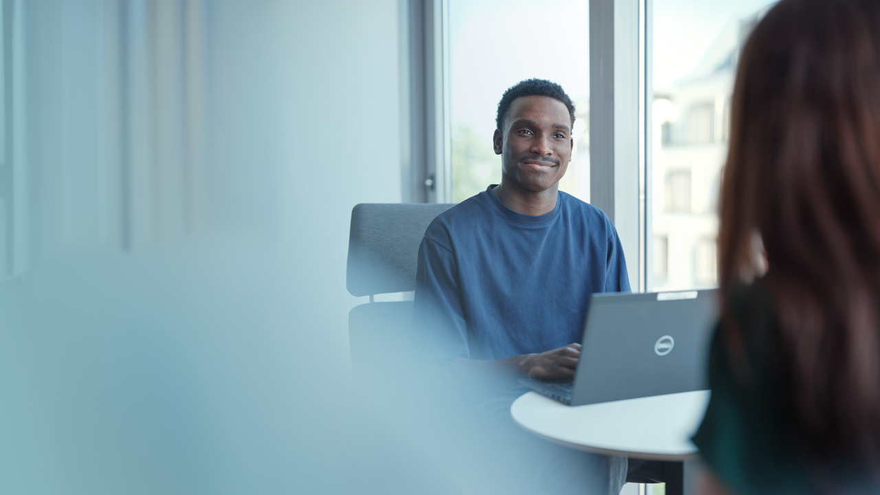 Un homme sourit tout en discutant avec une collègue. Il a son ordinateur portable. Nous devinons qu'ils sont en réunion.