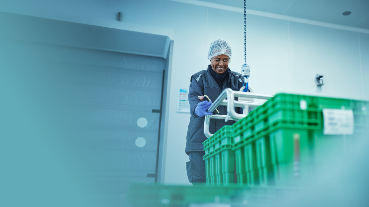 Une femme, très souriante, est en train porter une caisse de plasma avec un appareil d'aide au port de charge lourde. Elle porte une charlotte sur la tête et des gants violets.