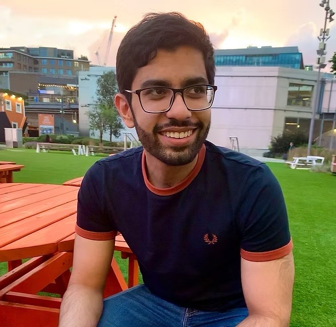 An Asian man with glasses and a beard smiles while sitting on a red bench in a grassy outdoor area with buildings in the background.