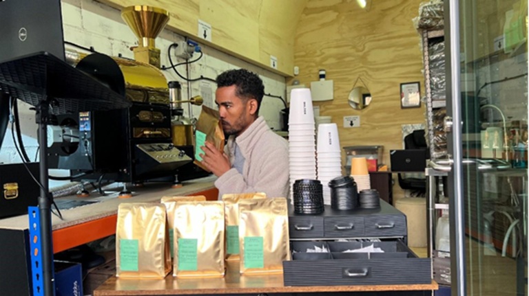 Man in a coffee shop smelling a bag of coffee beans, surrounded by stacked cups and bags of coffee on a counter.